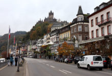 Die Moselpromande in Cochem. Links von der Straße fließt die Mosel, auf der rechten Seite finden sich Lokale, die zu gutem Essen und Wein einladen. Über der Promenade thront auf einem Hügel die Reichsburg von Cochem.