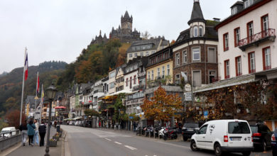 Die Moselpromande in Cochem. Links von der Straße fließt die Mosel, auf der rechten Seite finden sich Lokale, die zu gutem Essen und Wein einladen. Über der Promenade thront auf einem Hügel die Reichsburg von Cochem.