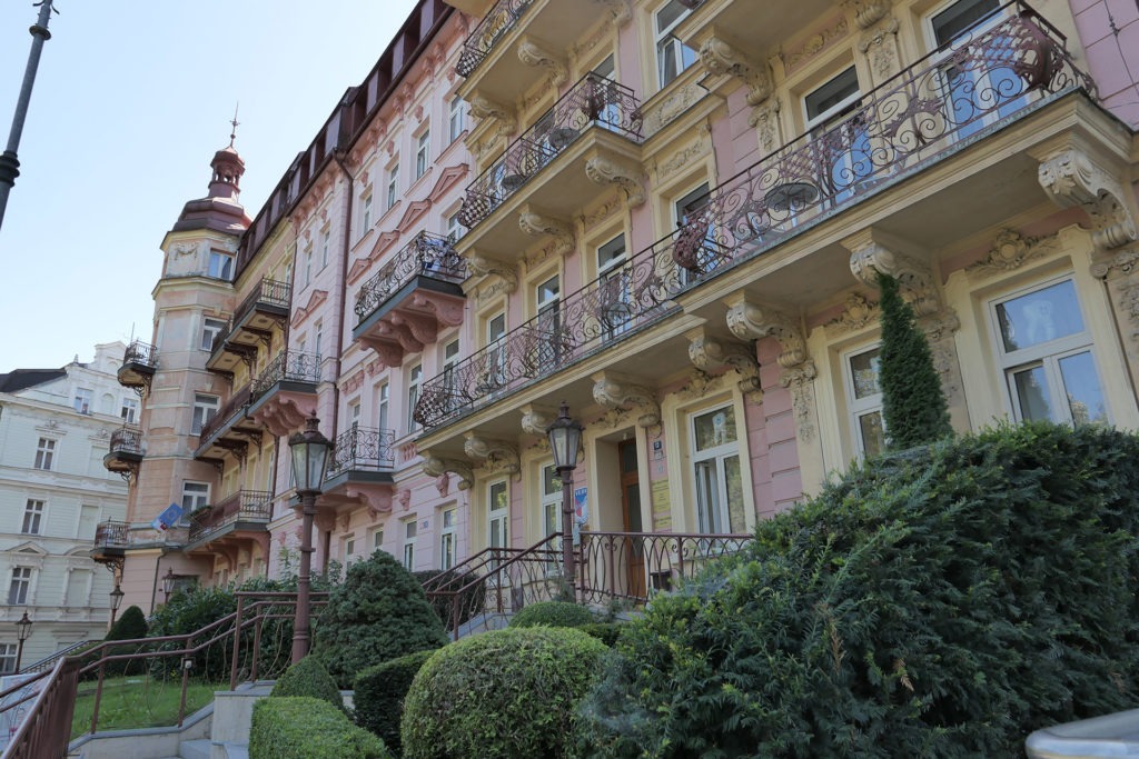 Das Bild zeigt drei Häuserfronten - allesamt Altbauten in unterschiedlichen Farben. Vor den Gebäuden stehen Büsche, die sehr gepflegt wirken. Das hinterste Gebäude hat an der linken Seite einen Turm.